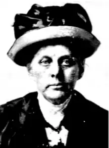A middle-aged white woman wearing a brimmed hat, a white blouse with a high collar, and a dark jacket