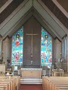 The interior of a building, with a vaulted ceiling supported by wood beams. The far end shows the back wall, which has a large wooden cross hung on a wall between two stained glass windows. An altar is visible before the wall, with a central path leading from it to the foreground, adjacent ot which are rows of pews.