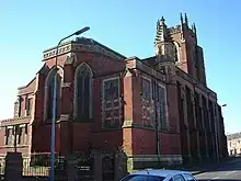 A broad church in brick with stone dressings, seen from the northeast, with a canted apse, a crocketted pinnacle, and in the distance a tower, also with crocketted pinnacles