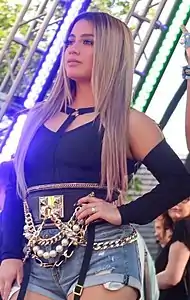Brooke, dressed in a black shirt with cut sleeves, denim shorts with gold chains, with her hand on her hip.