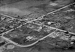 Aerial view showing wreckage, debris, and dismantled homes with farmland in the background