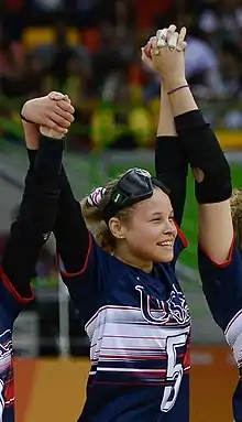 Dennis in her uniform, smiling with eye shades on her forehead. Her arms are raised and are holding the hands of two of her teammates.
