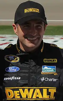 A man in his late thirties is wearing a black baseball cap and a black racing overall suit with sponsors logos.