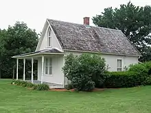 The exterior of the small home. Visible damage is seen near the front porch foundation, although restoration is also evident in metallic reinforcement of the shingled roof and chimney area.