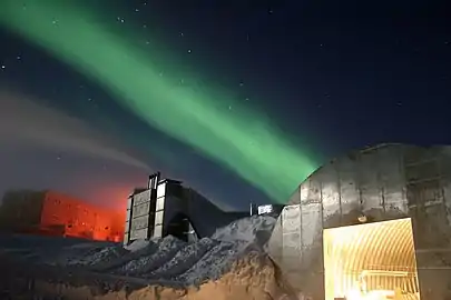 A low shot of a station at the South Pole taken at night. Nearest the front of the photo is a metal structure with a curved roof and a large, open door from which bright light emanates. Slightly further in the distance are two larger buildings. The sky above is a dark blue littered with stars and a green light present across the middle of the sky.