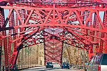 A red steel bridge arch is seen from a car going under the metalwork along the bridge deck