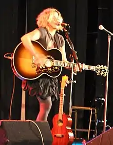 A woman with a guitar stands in front of a microphone stand