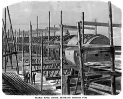 An engraving. A precarious structure of timber posts and beams supports a large iron cylinder with projecting flanges between segments. Three gentlemen stand in the foreground dwarfed by the structure and another sits on top astride one of the flanges.