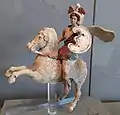 A female warrior is seated on the back of a rearing horse. She wears a one-shouldered tunic that flows behind her, a feathered helmet, a shield and jewelry, which are decorated with reds, browns, white, and gold. The horse's bridle and reins are similarly decorated.