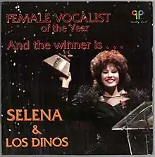 The cover of Selena's album "And the Winner Is..." features a photograph of Selena smiling broadly, wearing a black off-the-shoulder top, and proudly holding a Tejano Music Award. The title of the album is displayed in bold red capital letters above Selena's head, while Selena y Los Dinos appears in a larger orange font on the bottom left side.