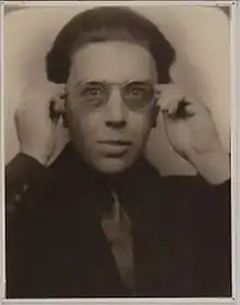 A sepia photograph of a man dressed in a suit. He is wearing a pair of semi-transparent sunglasses and has both hands raised to either side of his head. The photograph is surrounded by a white border.