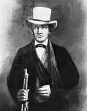 A man in a dark suit with a white hat. His left arm seems to be in a sling. In his right hand he grasps the barrel of a rifle.