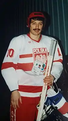 A male ice hockey player poses for a photo, visible from the knees up. He is dressed in ice hockey gear, wearing the national jersey of Poland, and while his helmet is on, he holds his gloves.