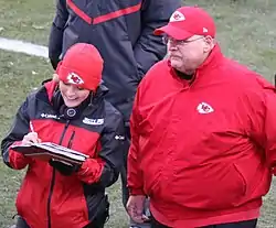 A reporter in a KCTV-branded jacket and writing in a notepad talking to Andy Reid on a football field