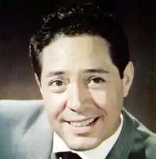 A dark-haired man in a light blue jacket, smiling slightly