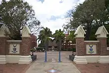 A brick wall with a gap in the middle. Through the gap one can see a statue at the bottom of a palm-lined walkway.