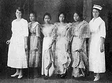 Six women standing in a row indoors. The first woman on the left is white, wearing a white dress. The second woman in Filipina, wearing a western-style dress; the next three women are also Filipinas, wearing "butterfly sleeve" formal dresses; the sixth woman is white, and tall, and wearing a white nurse's uniform and cap.