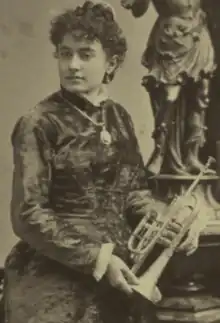 A white woman wearing a gown and a large pendant, holding a cornet or trumpet