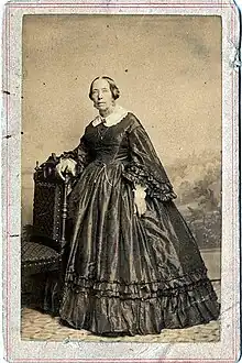 Photograph of a woman in a voluminous dress standing next to a chair