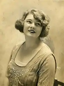 A sepia-toned photograph of a young Anne Roselle, smiling; her wavy hair is bobbed; she is wearing an embroidered dress with a low square neckline.