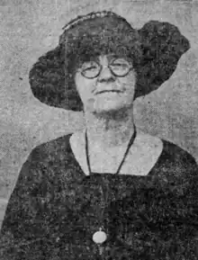 A white woman wearing a black brimmed hat, round eyeglasses, and a black dress