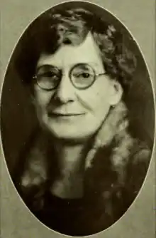 A nolder white woman with wavy hair, wearing round eyeglasses; her jacket or top has a high collar; in an oval frame