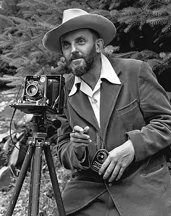 A photo of a bearded Ansel Adams with a camera on a tripod and a light meter in his hand.  Adams is wearing a dark jacket and a white shirt, and the open shirt collar is spread over the lapel of his jacket.  He is holding a cable release for the camera, and there is a rocky hillside behind him.  The photo was taken by J. Malcolm Greany and first appeared in the 1950 Yosemite Field School Yearbook.