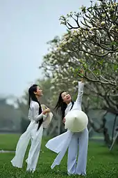 Photograph of two girls wearing a traditional Vietnamese white school uniform, the áo dài—both are holding the nón lá, a conical hat