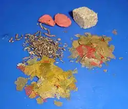 On a blue background, two large light red wafers at the upper left, a light gray compressed cube at the upper right, small brown pellets at the middle left, and, at the middle right and bottom, various yellow-green and red flakes