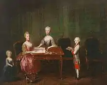 Four children around a table, a toddler in a blue dress on the left, a young teenager in pink seated and drawing, on her right a girl stands and helps her. A 6-year-old boy in a white-and-red military uniform is standing on the right with a book.