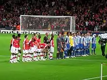 Two football teams and a refereeing team lining up in a football stadium pitch. The leftmost team wears red shirts and white shorts and socks, while the other wears blue shorts and socks and shirts with vertical blue-and-white stripes. The refereeing trio wears light gray equipment. A goal and a packed stand behind it are seen in the background.