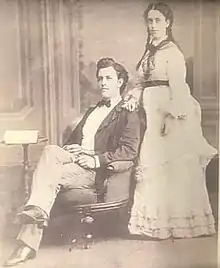 Black and white of a middle-aged man and woman. The man has dark hair, is seated in an armchair, wearing a dark-colored suit jacket, light-colored pants, white dress shirt and black bow tie. The woman is standing behind and to the side of the chair, wearing a full-length white dress with a dark-colored belt, and dark hair braided into two ponytails.