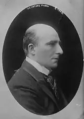 Right profile of white man with bald head except for a small amount of dark hair at the back and sides, clean shaven, with bushy eyebrows