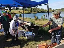 A man turns a spit with chochoca on it, while a woman brushes it, presumably with lard