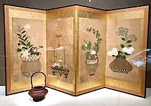 A small, squat flower basket, woven from dark brown bamboo, with a tall, arching handle, sits in front of a folding screen with a design of flower baskets on four panels.