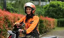A woman wearing orange jacket and helmet with "OK-JEK" written on them and riding a motorcycle