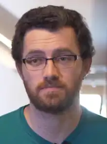 Austin Wintory speaking on video in 2019.  He is standing against a blurry office background, wearing a green shirt and glasses