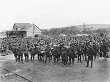 Black and white photo of a large group of men wearing military uniform standing in front of a small building. One rail of a railway track is visible in the foreground.