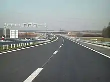 Six lane motorway, back side of a gantry supporting variable traffic signs is visible spanning the opposite direction carriageway