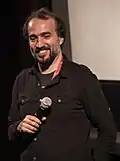 Ayat Najafi wearing a black shirt, holding a microphone in front of his belly, and smiling