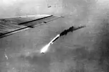 Black and white photo of flaming wreckage falling towards the ground. The wing of a plane is visible at the left-hand side of the photo.