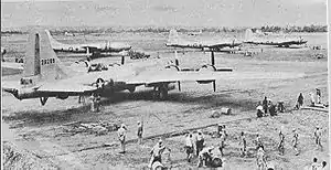 Four 4-engined World War II-era aircraft sitting on the ground at an airstrip. Groups of people are working near each aircraft.