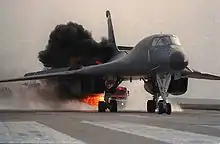 Black aircraft trailed by a column of black smoke and fire on the runway as fire trucks close in on the flame from behind