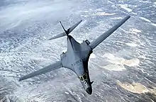 Top forward view of gray aircraft with wings swept forward banking right. Underneath are strips of white clouds and uninhabited terrain.