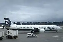 Right side view of an airplane taxiing on the tarmac, with several trucks in the foreground and to the left. In the background is a tree-covered hill and dark clouds.