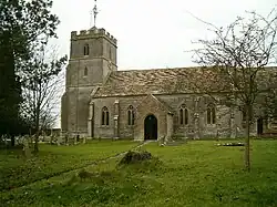 Brown stone building with square tower at the left hand end. In the foreground is a grass area with gravestones.