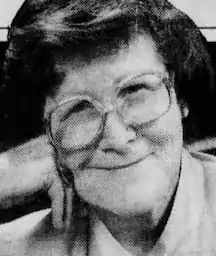 An older white woman, smiling, wearing glasses; her head is resting on her folded right hand.