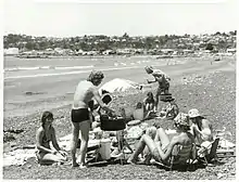 A group of people on a beach sitting around a barbecue grill.