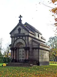 A reddish-brown stone building with a pointy roof, a cross on the front and a round-arched front door with the word "RICHMOND" carved into the stone above it, seen on an autumn day with filtered sunshine coming from the right and yellow leaves falling in front.