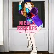 A red haired woman wearing a colored dress sitting on a window. In front of her, it is written "NICOLA ROBERTS BEAT OF MY DRUM" in pink.